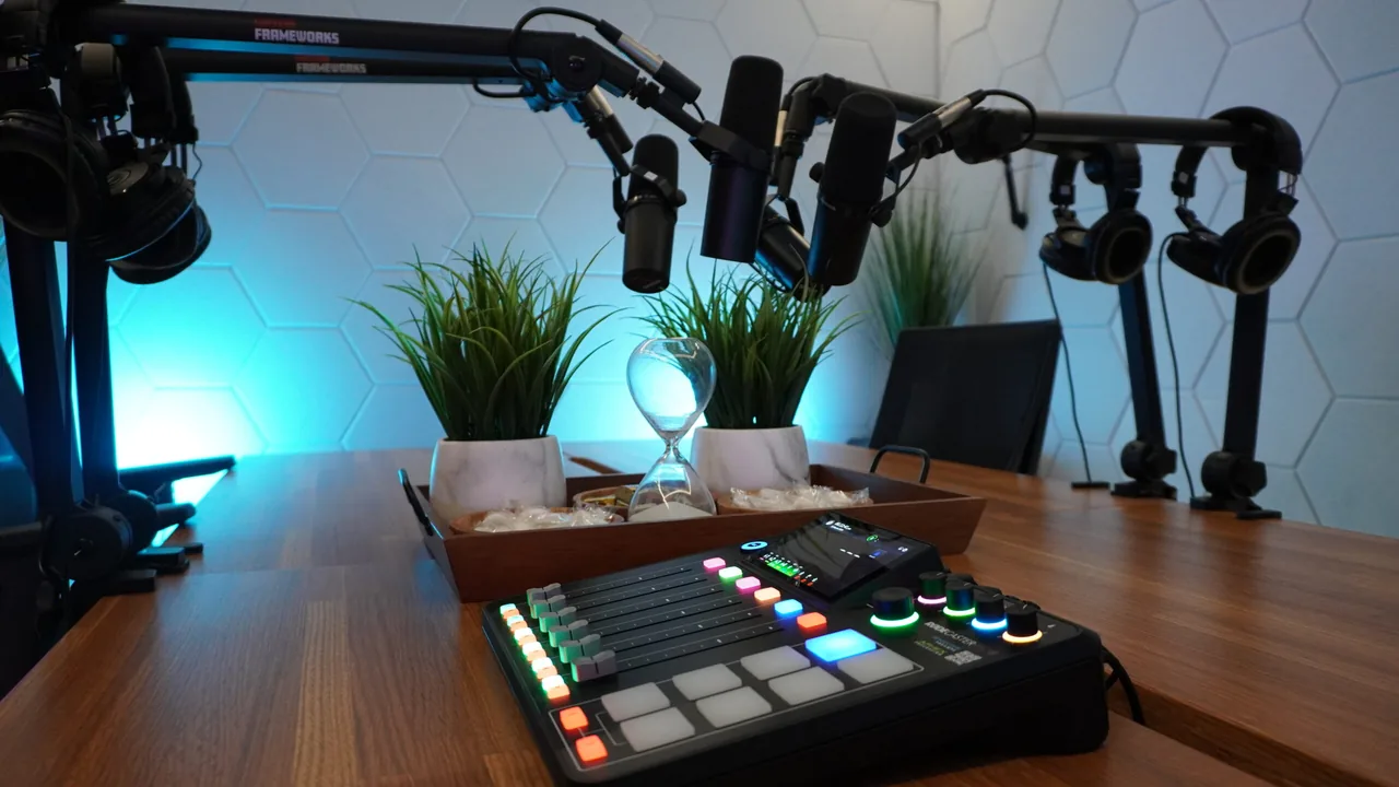 Hourglass between two potted plants on the four-mic podcast table at Podcast Studio San Antonio, with a RODECaster Pro II console in front and blue light on a hexagon acoustic wall